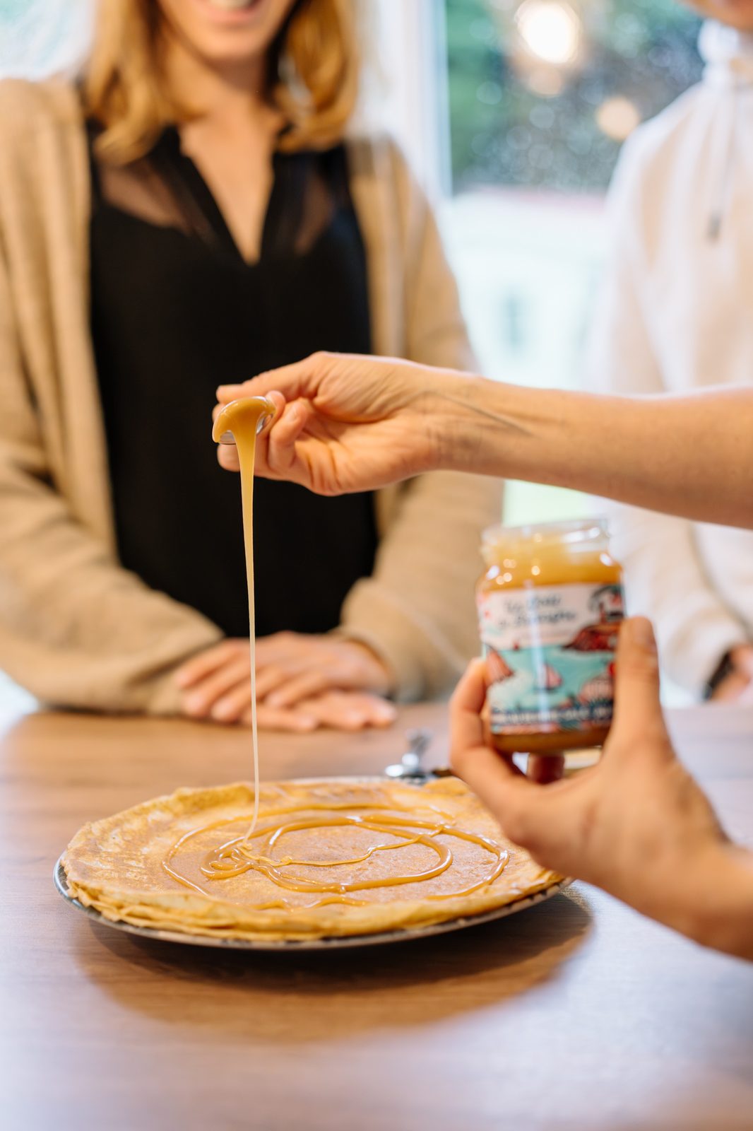 Une cuillère de caramel au cidre s'étire en filet sur une crêpe, à côté du pot Un Goût de Bretagne.